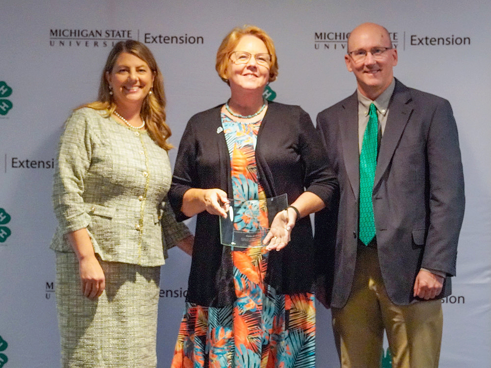 Three individuals at a backdrop. The center person holds an award.